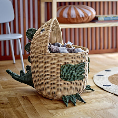 Eddi Dinosaur Rattan Basket in natural with green woven spines, arms and clawed feet, shown holding blankets on a wooden floor in a living room setting.