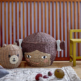 Lill Basket with Lid in brown Bankuan grass, woven as a smiling face with two side bunches, shown beside a bear-shaped rattan basket in a nursery setting.