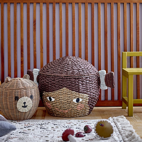 Lill Basket with Lid in brown Bankuan grass, woven as a smiling face with two side bunches, shown beside a bear-shaped rattan basket in a nursery setting.