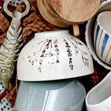 Bea Floral Stoneware Bowl 11.5cm in beige, shown upturned with fine sprig floral motif, glossy finish, among mixed ceramics and wooden items.