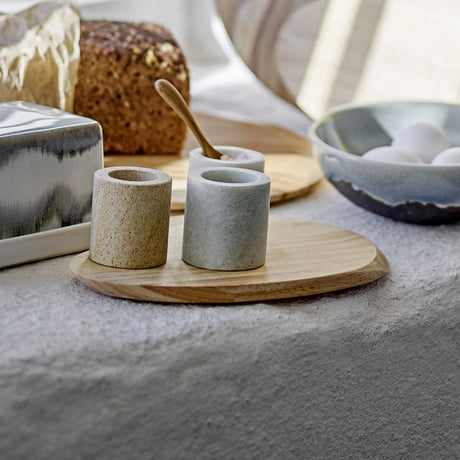 Oval natural wood pebble-shaped chopping board from the Gong set, holding stone egg cups and a spoon beside bread and a ceramic bowl of eggs.