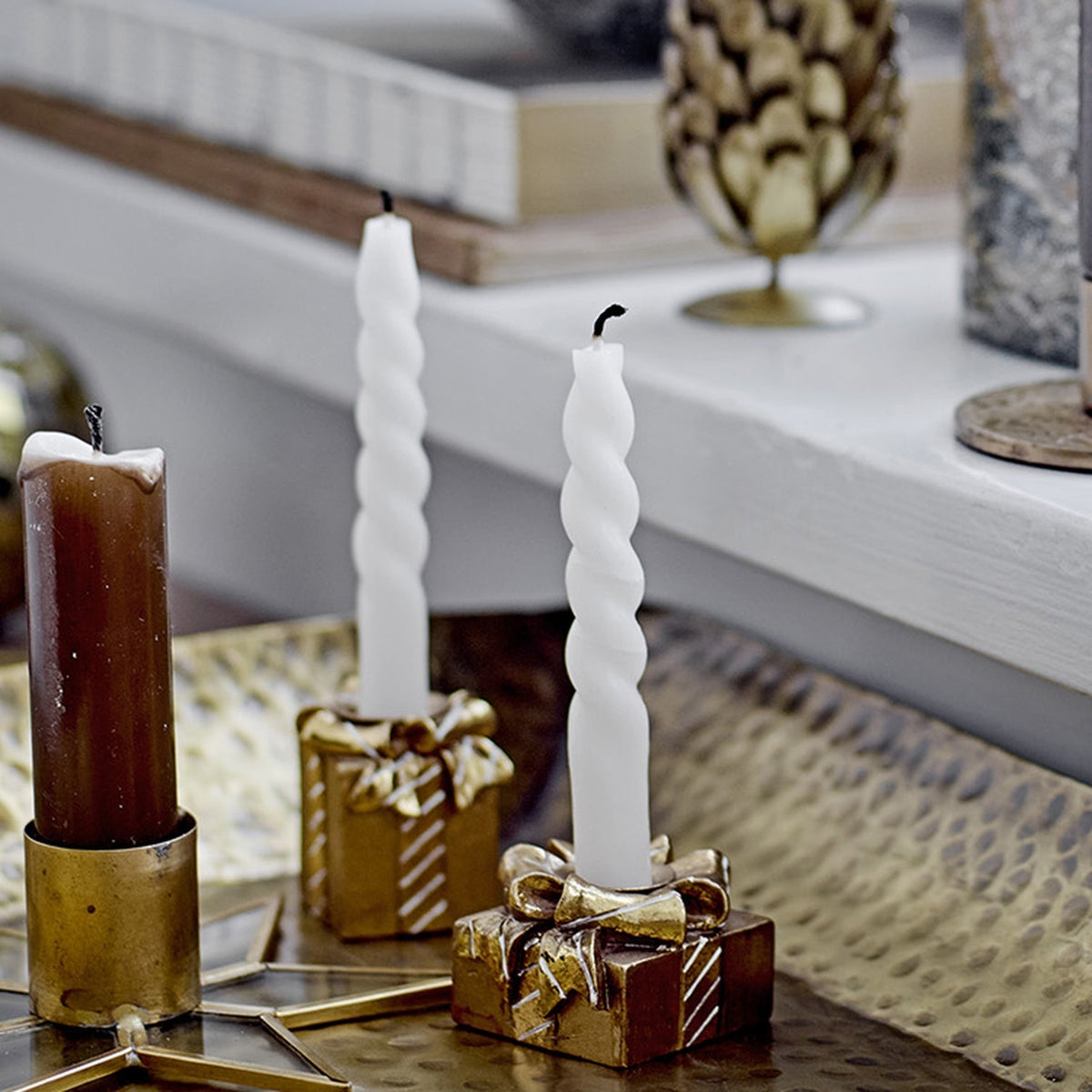 Two white twist candles from the Twist Candles Pack of 12 in slim spiral paraffin wax, displayed upright in gold gift-box candle holders beside a brown pillar candle.