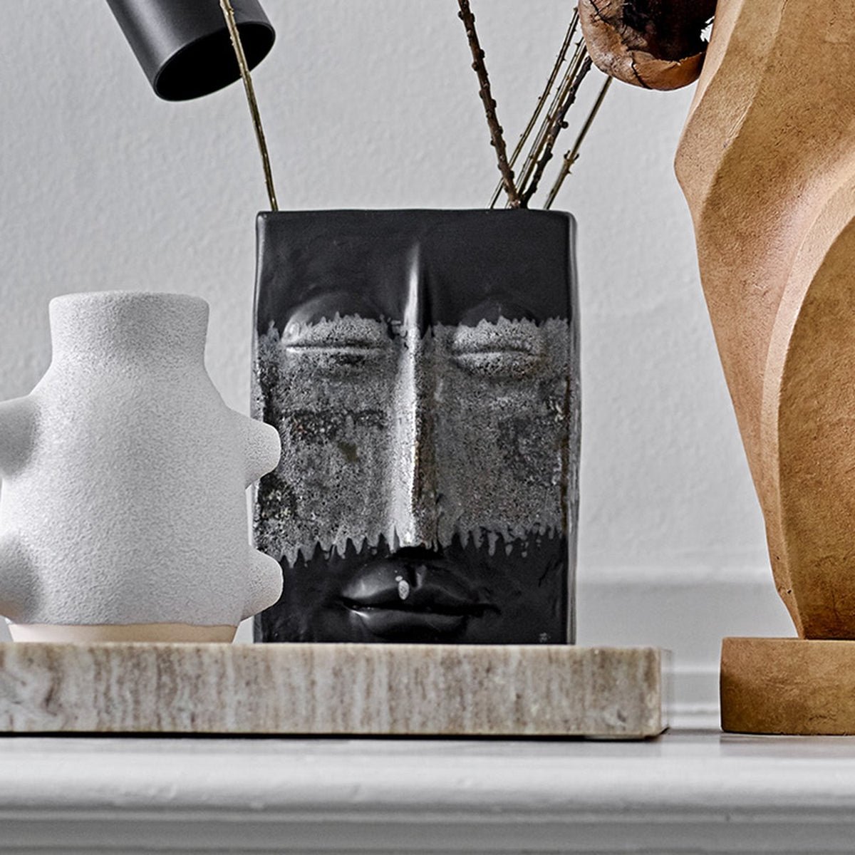 Nasru Stoneware Vase 22cm in Black, a rectangular ceramic vase moulded with a calm closed-eyed face and speckled glaze, shown with twigs on a shelf beside white and wood decor pieces.