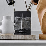 Nasru Stoneware Vase 22cm in Black, a rectangular ceramic vase moulded with a calm closed-eyed face and speckled glaze, shown with twigs on a shelf beside white and wood decor pieces.