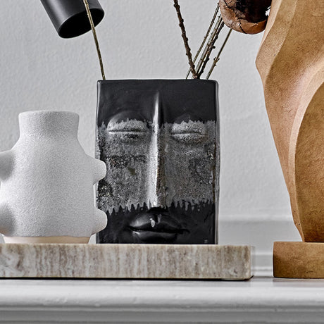 Nasru Stoneware Vase 22cm in Black, a rectangular ceramic vase moulded with a calm closed-eyed face and speckled glaze, shown with twigs on a shelf beside white and wood decor pieces.