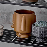 Guxim Stoneware Plant Pot in brown with a face-shaped design, tapering nose, blocky ears and ring foot, shown on a shelf beside a succulent planter.