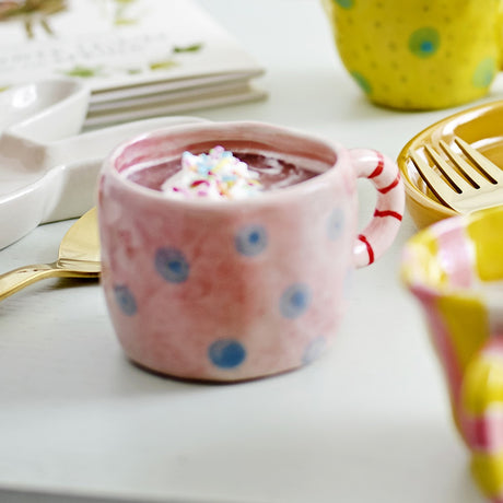 Nini Dotted Stoneware Cup 100ml in pink with blue polka dots and a red striped handle, shown filled with dessert and set on a table with a gold spoon.