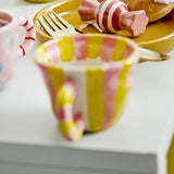 Nini Yellow Striped Stoneware Cup 110ml, a small handle-equipped ceramic cup with yellow and pink vertical stripes and a candy-cane style handle, shown beside a matching pink mug and striped plate.