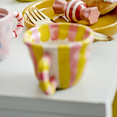 Nini Yellow Striped Stoneware Cup 110ml, a small handle-equipped ceramic cup with yellow and pink vertical stripes and a candy-cane style handle, shown beside a matching pink mug and striped plate.