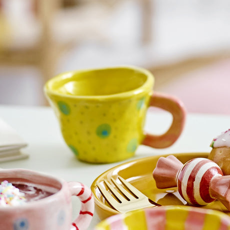 Nini Stoneware Cup 100ml in yellow with hand-painted spots and a pink handle, shown on a table beside a matching plate and playful tableware accents.