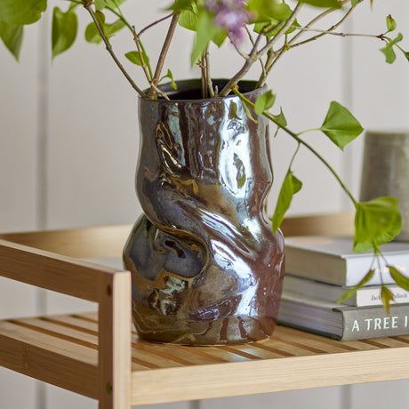 Apio Stoneware Vase 22.5cm in grey with a twisted, pinched ceramic body and glossy glaze, shown with foliage on a wooden shelf beside stacked books.