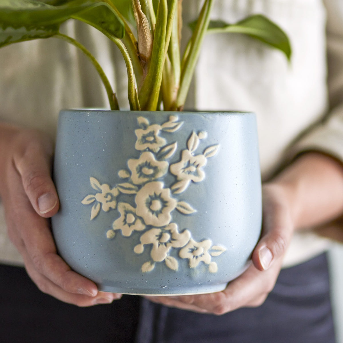 Betino Floral Stoneware Plant Pot in blue, held in two hands, showing a raised white floral spray on a speckled blue ceramic planter with a leafy plant inside.