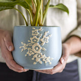 Betino Floral Stoneware Plant Pot in blue, held in two hands, showing a raised white floral spray on a speckled blue ceramic planter with a leafy plant inside.