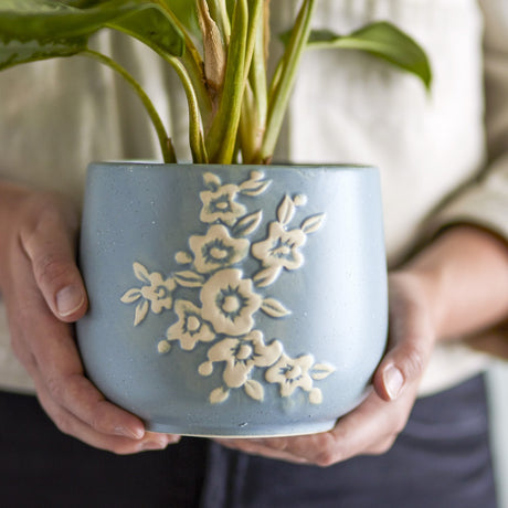 Betino Floral Stoneware Plant Pot in blue, held in two hands, showing a raised white floral spray on a speckled blue ceramic planter with a leafy plant inside.