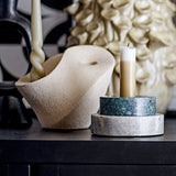 Brent stoneware candle holder in beige with sculptural curved body and sandy speckled finish, shown beside a marble pillar candle stand on a dark surface.