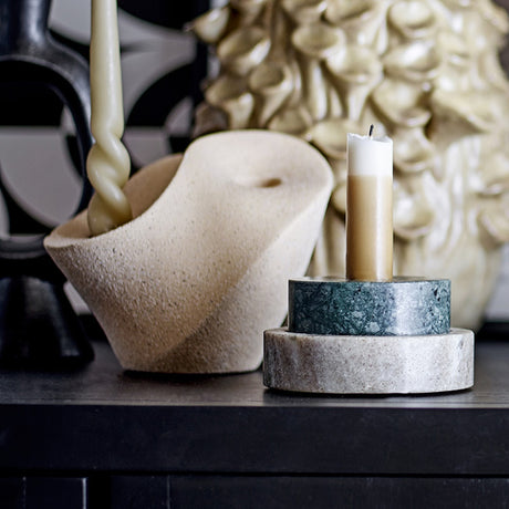 Brent stoneware candle holder in beige with sculptural curved body and sandy speckled finish, shown beside a marble pillar candle stand on a dark surface.