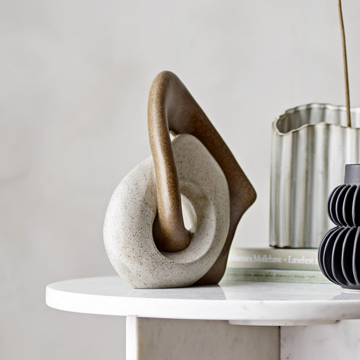 Esad Resin Ornament in brown, a sculpted interlocking loop design with a speckled white base, displayed on a marble table beside books and vases.