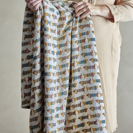 Charlie Cotton Throw in Multi being held up, a cotton-blend children's throw patterned with rows of striped sausage dogs in mustard, rust, teal and grey.