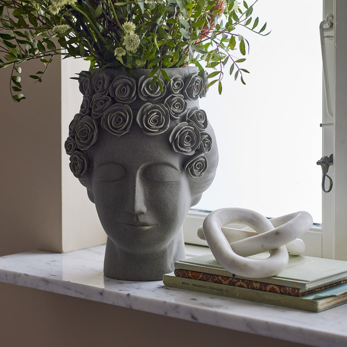 Akira Stoneware Vase 30cm in grey, shaped as a woman's head with closed eyes and a raised rose wreath crown, holding foliage on a marble windowsill beside books and a marble knot ornament.