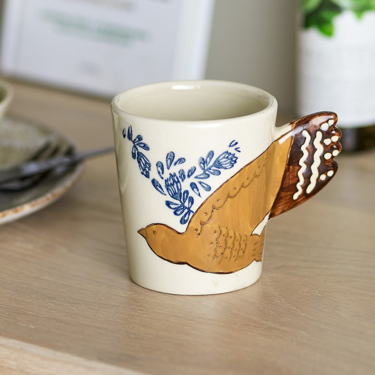 Hezha Stoneware Cup 260ml in cream, hand-painted with a mustard bird in flight forming the handle through its fanned tail, with blue floral sprigs, shown on a wooden table.