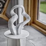 Rouyi Stoneware Ornament in grey, a sculptural abstract figurine with twisting looped arms, displayed on a round marble pedestal in a living room setting.