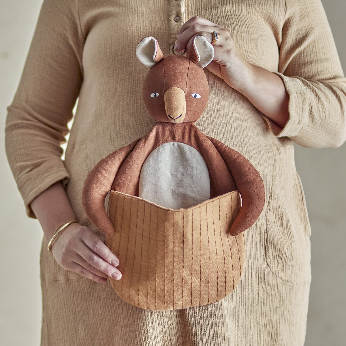 Toto Linen Wall Basket in brown, a kangaroo-shaped fabric storage piece with a quilted front pouch, held against a person's beige dress in a nursery setting.