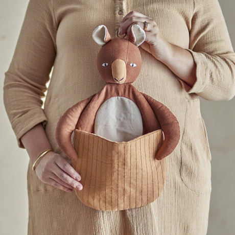 Toto Linen Wall Basket in brown, a kangaroo-shaped fabric storage piece with a quilted front pouch, held against a person's beige dress in a nursery setting.