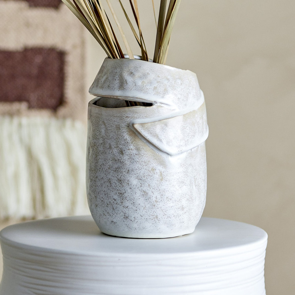 Abeera Stoneware Vase 20cm in beige, textured ceramic with a folded, overlapping band design, shown with dried reeds on a white pedestal against a neutral background.
