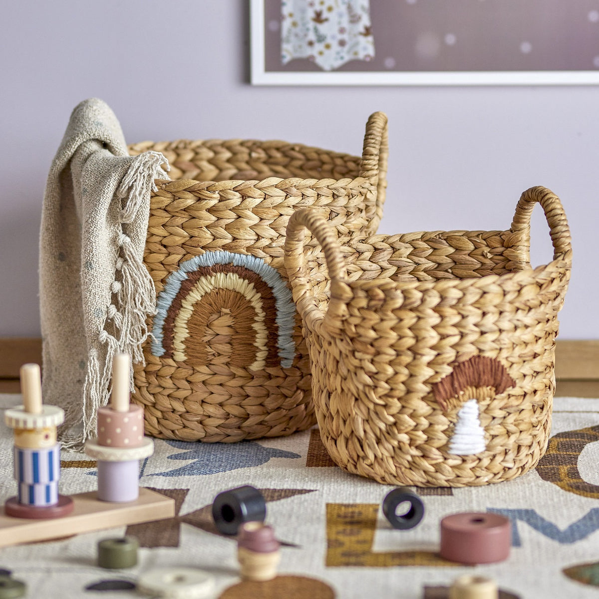 Agnes Water Hyacinth Baskets Set of 2 in beige, round woven storage baskets with handles, one stitched with a rainbow motif and the smaller with a mushroom design, shown in a nursery setting.