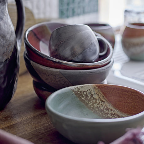 Paula Stoneware Bowl 16cm in green, shown with a diagonal terracotta band and reactive glaze, stacked with other ceramic bowls and a mug on a wooden surface.