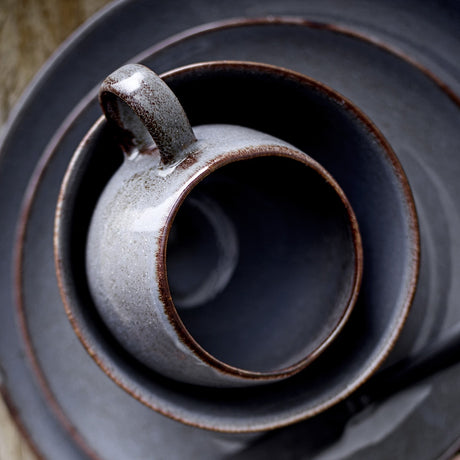 Top-down view of the Sandrine Stoneware Mug 500ml in grey, showing the speckled reactive glaze and loop handle nested inside matching stoneware plates.