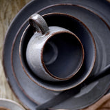 Sandrine Stoneware Cup in grey, round-bellied with a loop handle and speckled reactive glaze, shown nested in matching dark grey saucers from above.