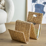 Dex Water Hyacinth Magazine Rack in Natural, woven zigzag design with two open slots holding a magazine, shown beside a boucle chair on a wooden floor.