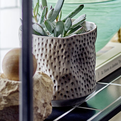 Sayah Stoneware Plant Pot in grey with dimpled texture and narrow foot, holding a succulent on a dark shelf with decorative stones nearby.