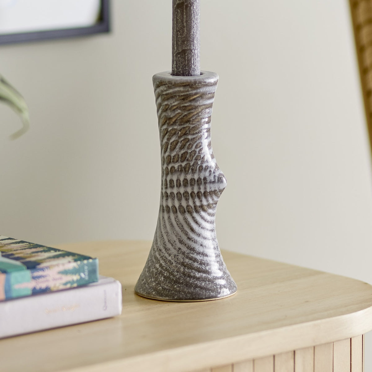 Raheem Stoneware Candle Holder in grey, 17cm hourglass shape with dimpled ridge texture and a small knot detail, shown on a wooden sideboard beside stacked books.