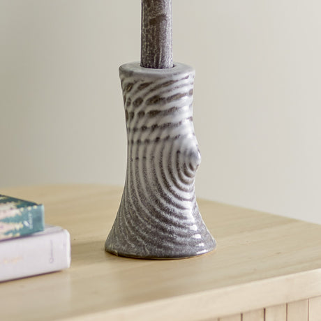 Raheem Stoneware Candle Holder 13cm in grey, a ceramic taper candle holder with a tapering, wood-like ridged body and raised knot detail, holding a candle on a wooden surface beside books.