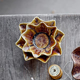 Dinah Flower Stoneware Tealight Holder in brown, star-shaped layered petal design with a round central well, shown on a wooden surface beside a thread spool.