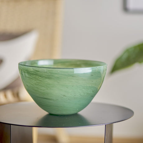 Esmaralda Glass Bowl 25cm in Green, a round glass bowl with swirled streaks and a wide thin rim, shown on a dark side table in a living room setting.