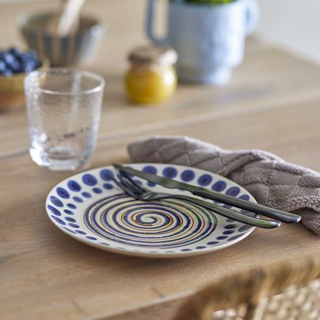 Shama Dotted Stoneware Side Plate in multicoloured with hand-painted spiral centre and dotted border, set on a wooden table with a knife and fork, glass, and napkin.