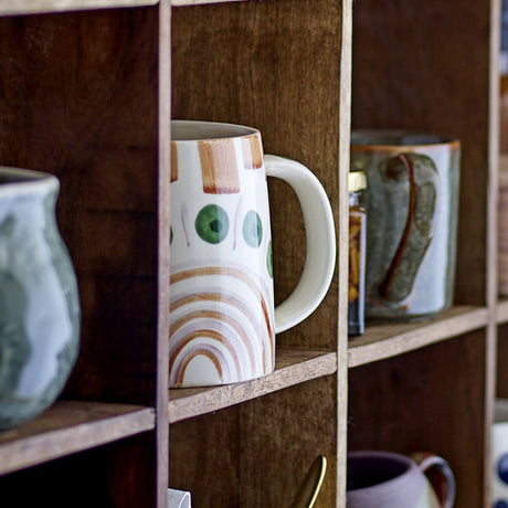 Shama Stoneware Mug 515ml in Multi, hand-decorated with rainbow and dot motifs, white body and rounded handle, shown on a wooden shelf among other ceramics.