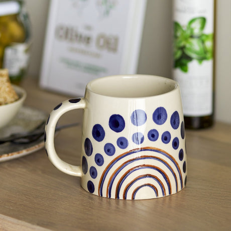 Shama Dotted Stoneware Mug 515ml in cream with hand-painted navy dot rings and brown concentric arches, shown on a wooden surface in a kitchen setting.