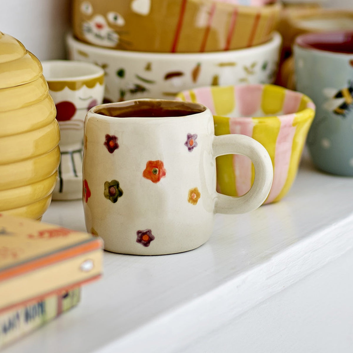 Addy Stoneware Cup 130ml in cream, a small rounded ceramic cup with scattered floral decoration and a loop handle, shown on a shelf among patterned bowls and cups.