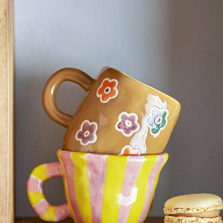 Addy Stoneware Cup 130ml in brown, glossy ceramic with hand-painted flower decals and a round loop handle, stacked atop a pink and yellow striped cup on a shelf.