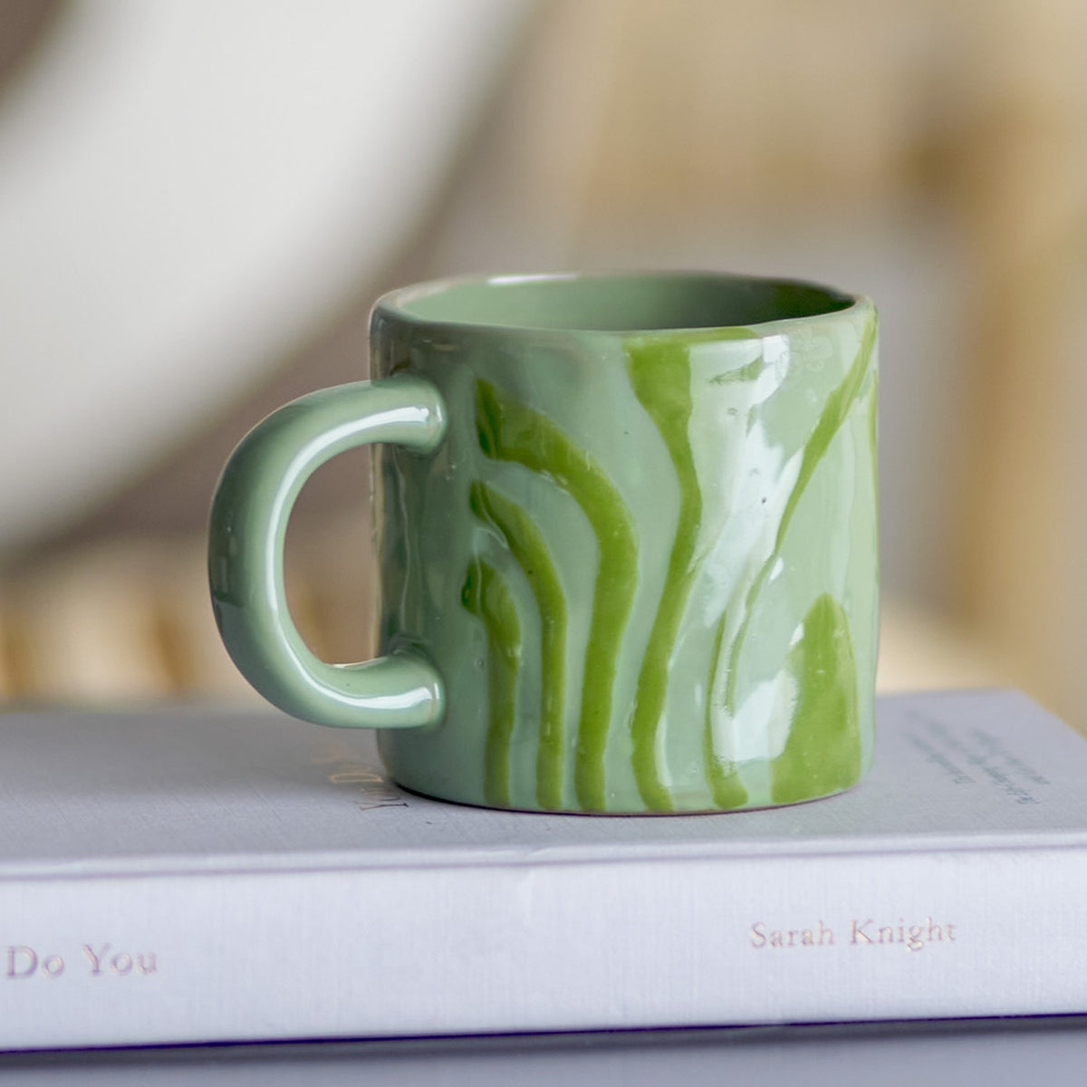 Ninka Stoneware Cup in green with glossy glaze and bold flowing stripes, rounded loop handle, shown atop a stacked book in a home setting.