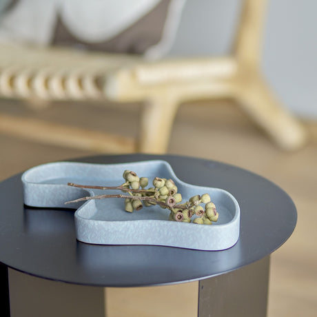 Lakey Stoneware Tray in blue with a freeform curved outline and low rim, shown on a black round side table holding dried botanical stems.