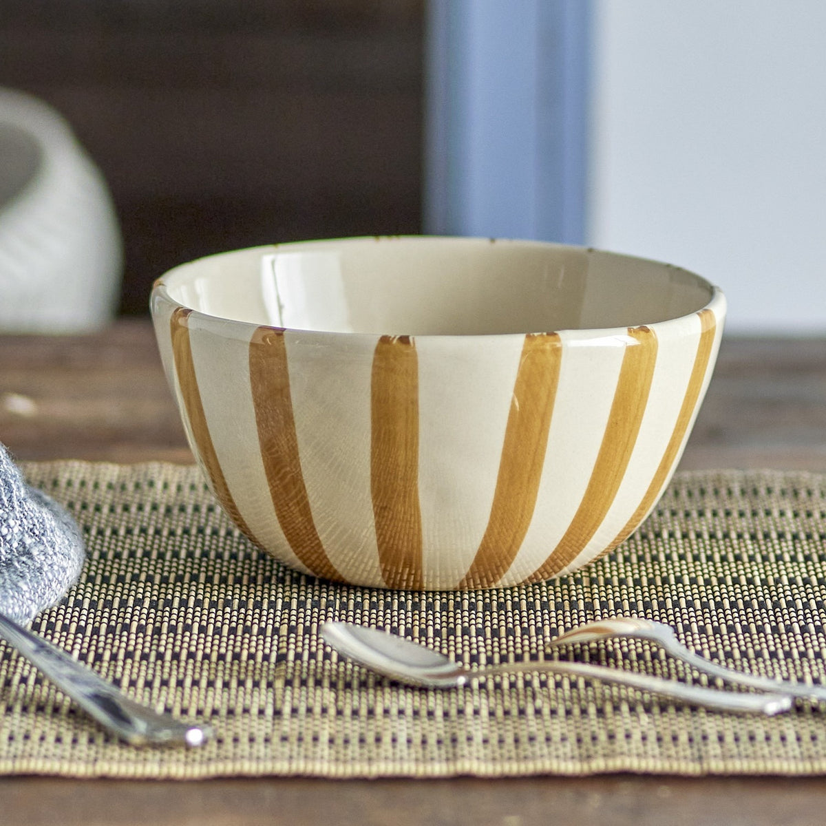 Begonia Striped Stoneware Bowl 14cm in Brown, a cream ceramic bowl with hand-painted brown vertical stripes, shown on a woven placemat beside cutlery in a dining setting.