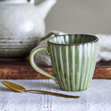 Latina Fluted Stoneware Cup 220ml in green with ridged body and loop handle, shown beside a gold teaspoon with a ceramic teapot in the background.