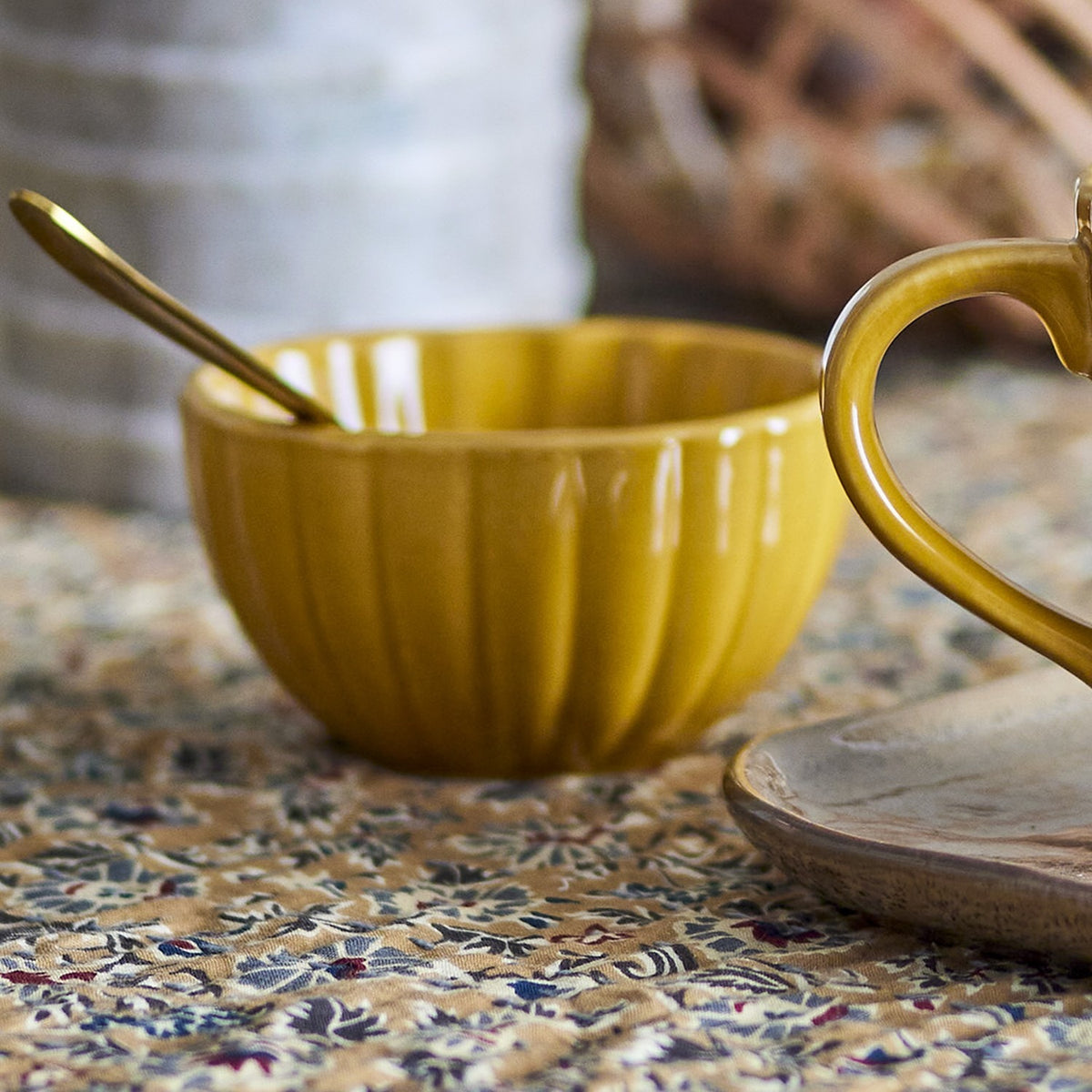 Latina Stoneware Bowl 12cm in yellow, round with fluted vertical ribs and a glossy ceramic finish, shown with a gold spoon on a patterned tablecloth.