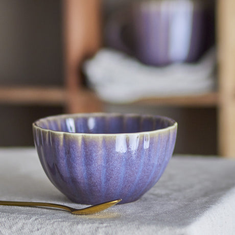 Latina Stoneware Bowl 12cm in purple, fluted sides with a glossy ceramic glaze, shown on a table with a brass-handled spoon beside it.