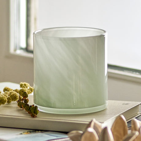 Sakkara Glass Tealight Holder in green, a straight-sided cylindrical glass votive, shown on a book beside dried flowers near a sunlit window.